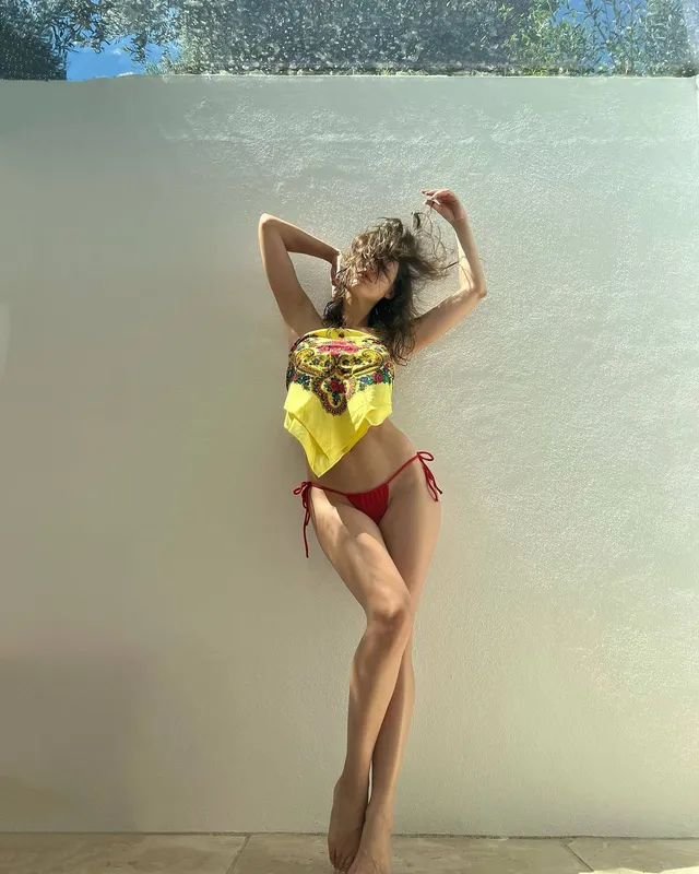 Úrsula Corberó standing barefoot by a wall in a colorful top and red bikini bottoms.