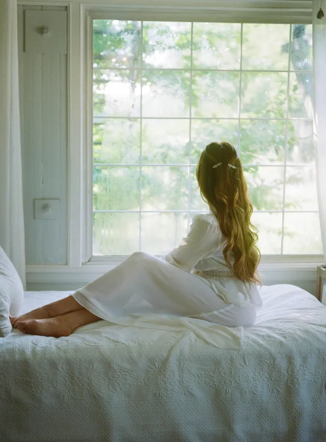 Elizabeth Gillies sitting on a bed beside a window.
