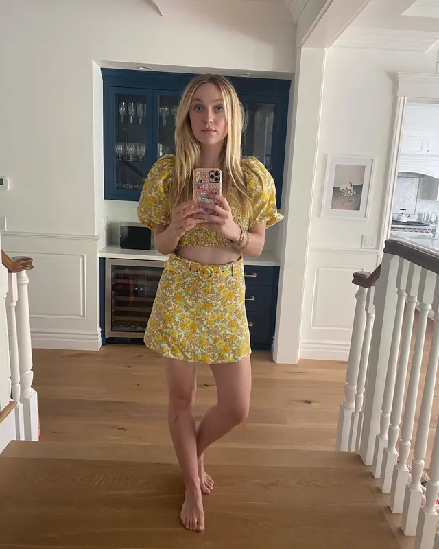 Dakota Fanning standing barefoot indoors in a yellow outfit.