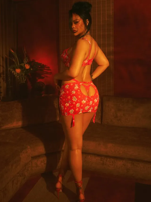 Rihanna posing in a red outfit and lace-up heels beside a sofa.