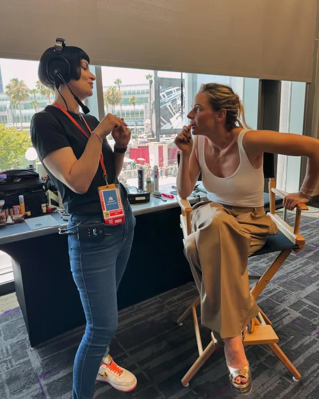 Rebecca Ferguson sitting in a makeup chair in a white top, tan trousers and gold heels.