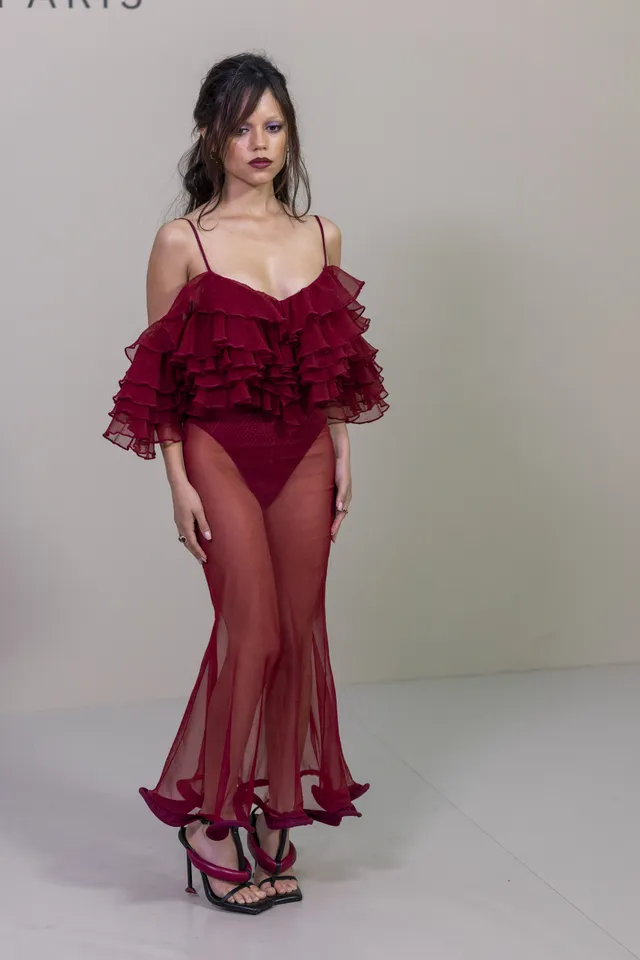 Jenna Ortega posing against a pale backdrop in a burgundy gown and heels.