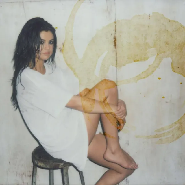 Selena Gomez sitting barefoot on a stool wearing a white shirt.
