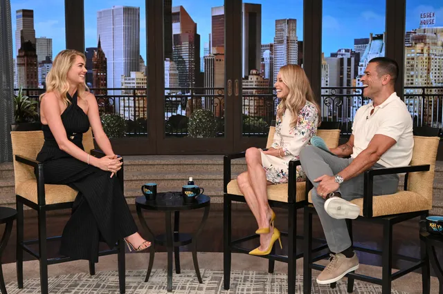Kate Upton sitting in a black outfit and heels during an interview.
