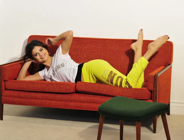 Selena Gomez reclining on a red sofa with bare feet raised.