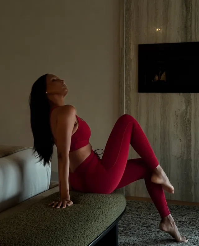 Jasmine Tookes stretching barefoot on a bench in a burgundy exercise outfit.