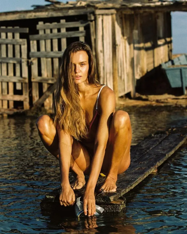 Josephine Skriver crouching barefoot on a wooden platform above water.