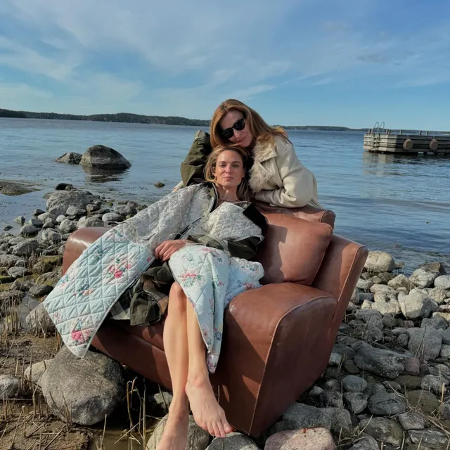 Rebecca Ferguson sitting barefoot on an armchair beside a companion by the sea.