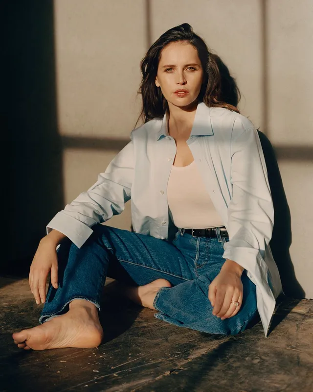 Felicity Jones sitting on a studio floor in jeans and a pale shirt.