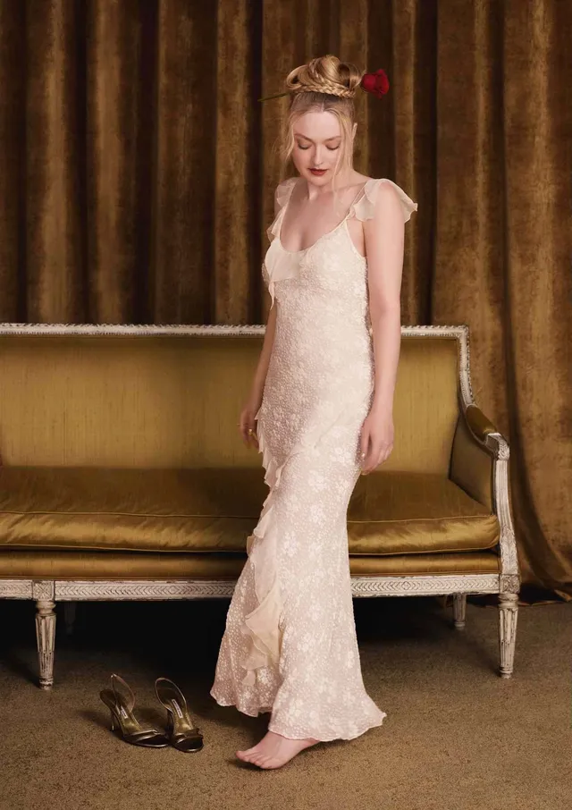 Dakota Fanning standing barefoot beside a sofa in a pale dress.