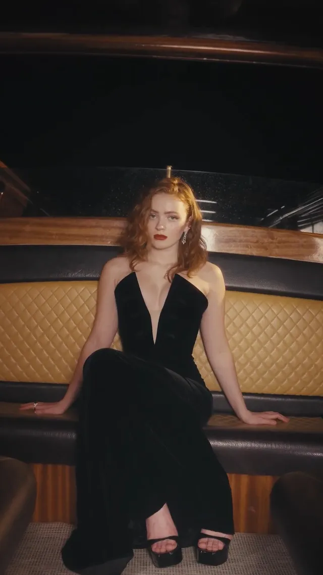 Sadie Sink sitting in a black dress and heels in a dim booth.