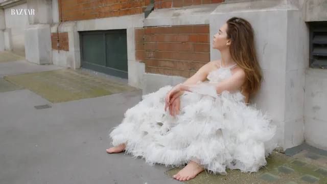 Emilia Clarke sitting barefoot beside a wall in a feathered dress.