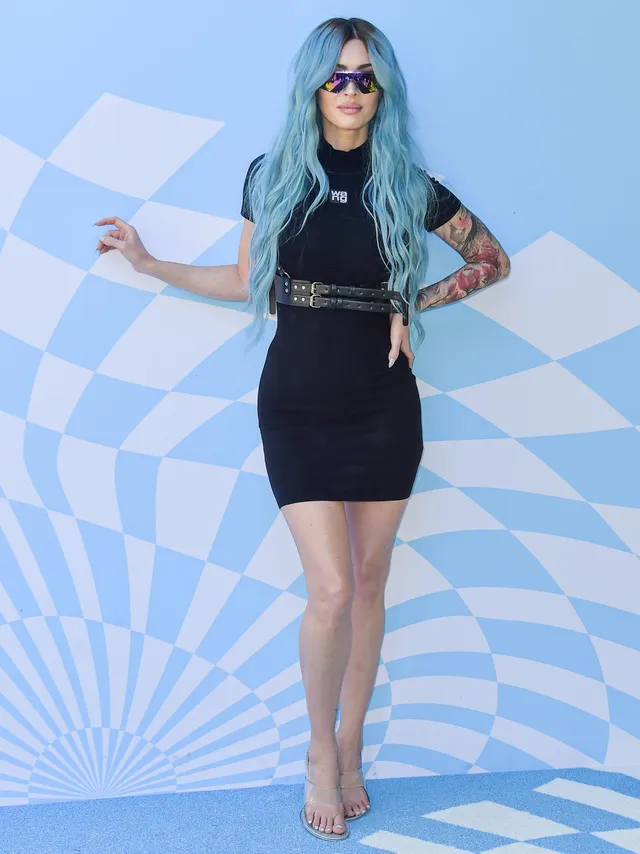 Megan Fox standing in a black mini dress and pale heels at an event.