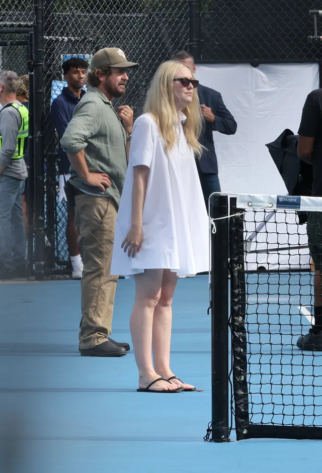 Dakota Fanning standing at a tennis court in a white dress and sandals.