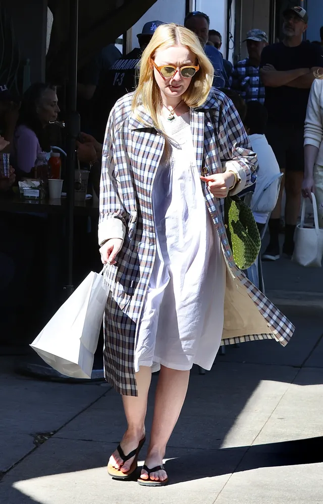 Dakota Fanning walking outdoors in a white dress and checked coat.