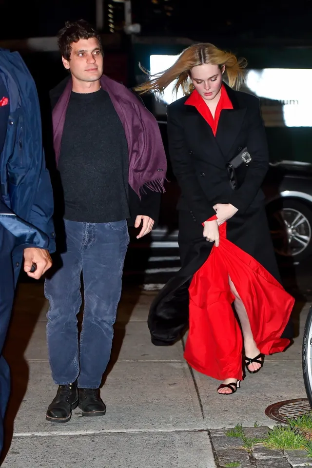 Elle Fanning walking outdoors in a black coat over a red dress.