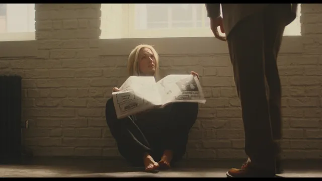 Sarah Pidgeon sitting barefoot on a floor while reading a newspaper.