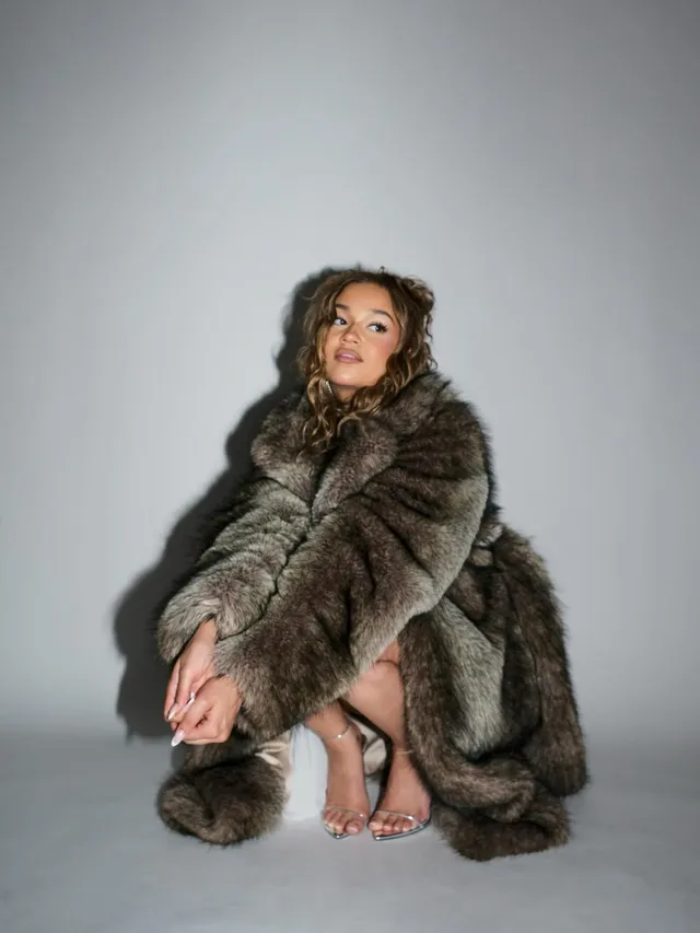 Madison Bailey crouching in a fur coat and clear heels in a studio.