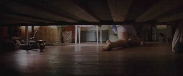Chloë Grace Moretz with bare feet visible beneath a bed on a wooden floor.