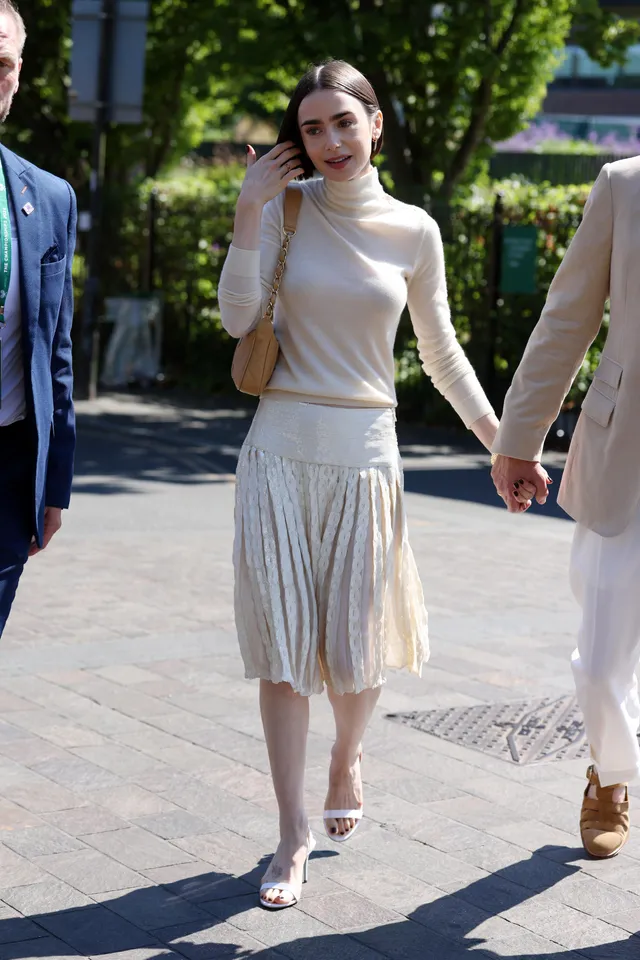 Lily Collins walking outside in a pleated skirt and pale heels.