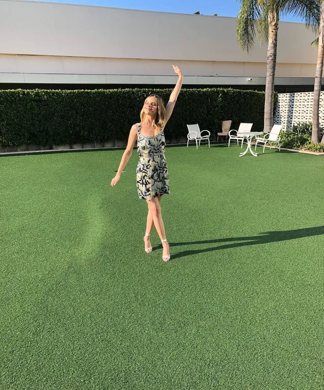 Halston Sage posing on a lawn in a floral dress and light heels.