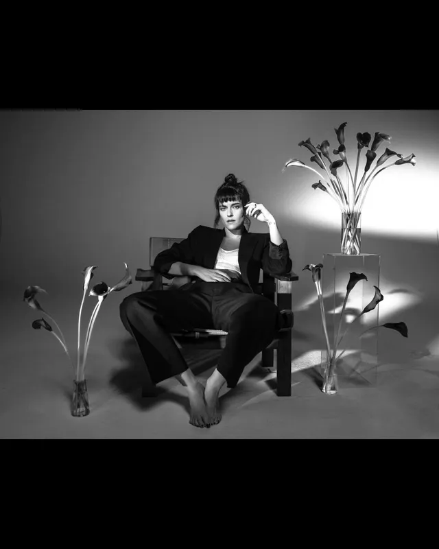 Kristen Stewart sitting barefoot on a chair in a monochrome studio scene.