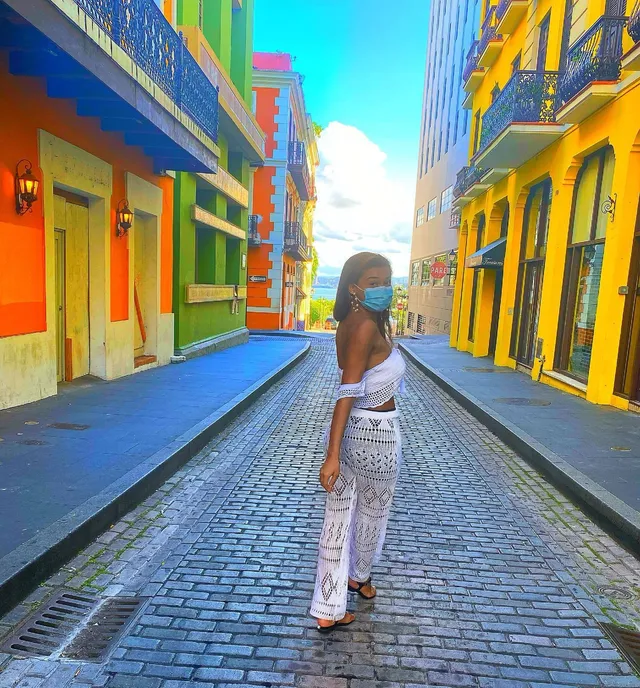 Sierra Capri standing on a colorful street in a white outfit and flat sandals.