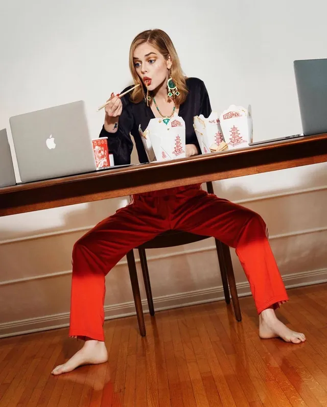 Samara Weaving sitting barefoot at a table in red trousers.