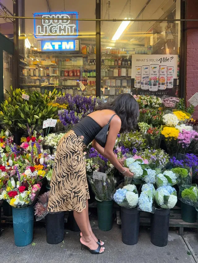 Greta Onieogou looking at flowers outside a shop in a patterned skirt and flat sandals.