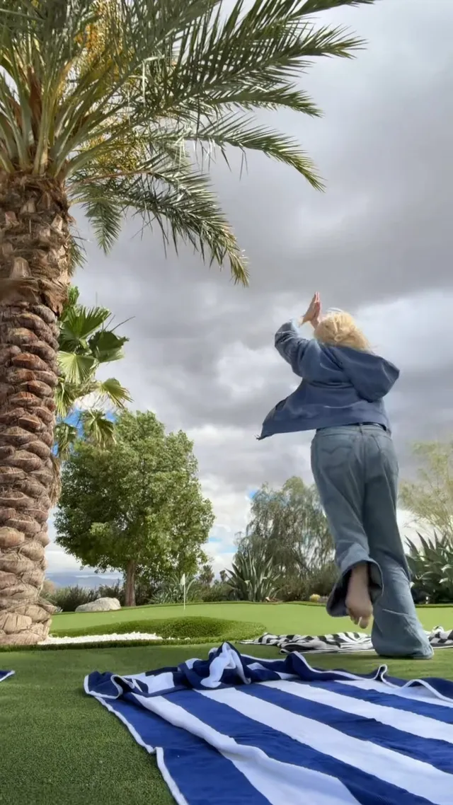 Rosé dancing barefoot on a lawn in jeans and a blue jacket, photographed from behind.