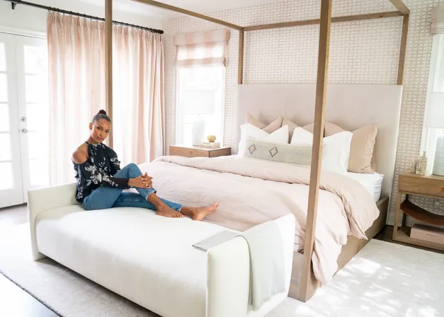 Jasmine Tookes sitting barefoot on a bed in a bright bedroom.