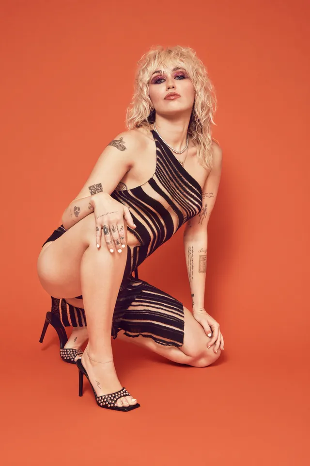 Miley Cyrus crouching in a striped dress and black heels for a studio portrait.