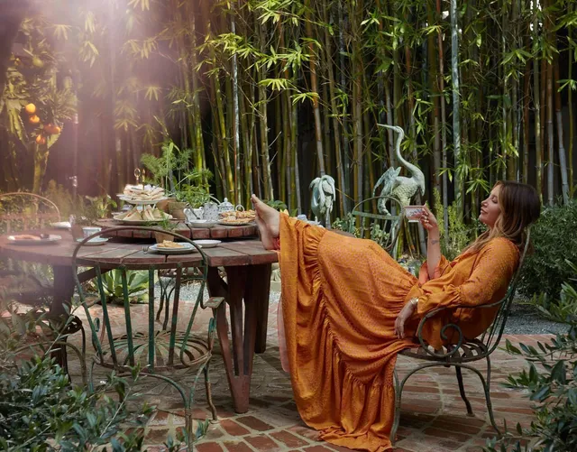 Ashley Tisdale reclining barefoot in a garden chair wearing a long orange dress.