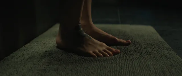 Dove Cameron standing barefoot on a dark upholstered surface.