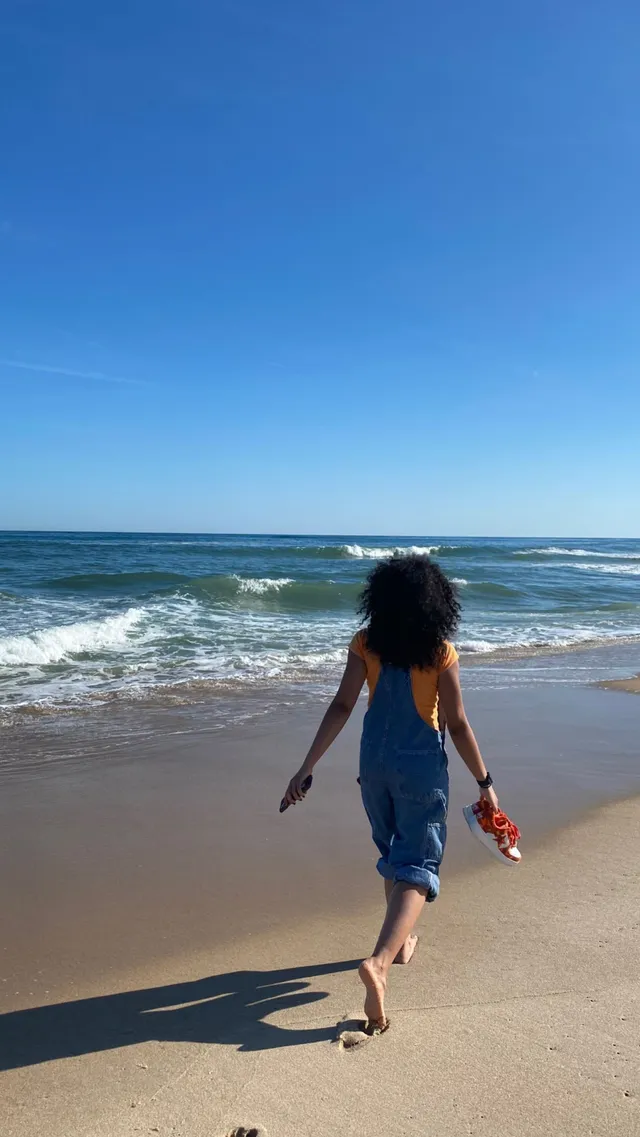 Whitney Peak walking barefoot on a beach in a blue outfit.