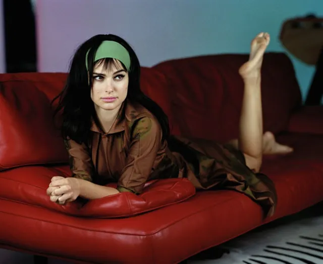 Natalie Portman reclining barefoot on a red sofa with one foot raised.