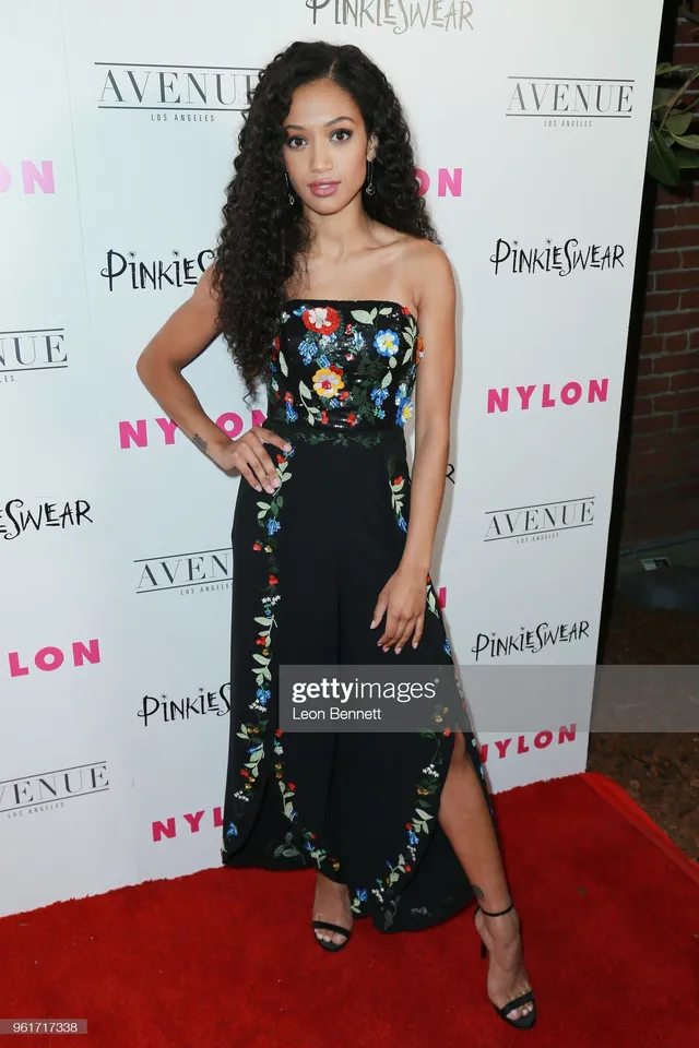 Samantha Logan posing in a black floral dress and heels before an event backdrop.
