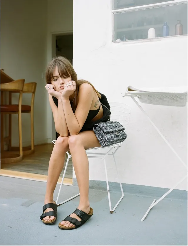 Ella Purnell sitting on a chair in flat black sandals.