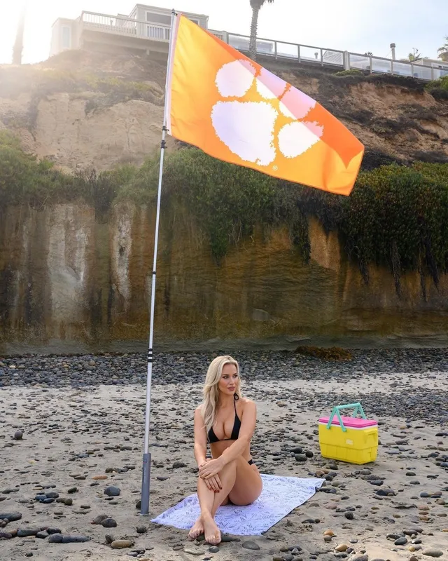 Paige Spiranac sitting barefoot on a towel beside a lakeside golf flag.