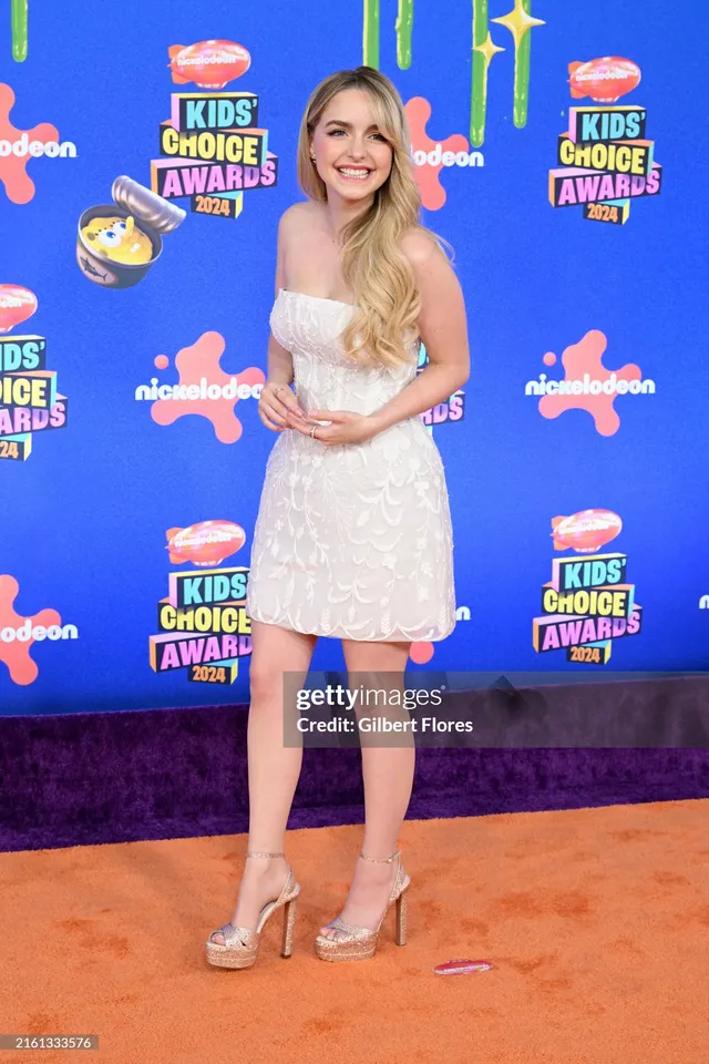 Mckenna Grace standing on an awards carpet in a short pale dress and metallic heels.