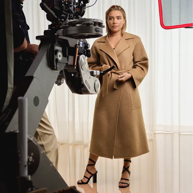 Florence Pugh standing beside a camera in a tan coat and black heels.