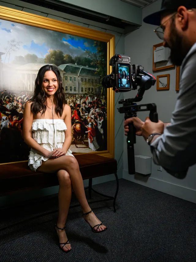 Olivia Rodrigo: seated in a white dress and heels while being filmed.