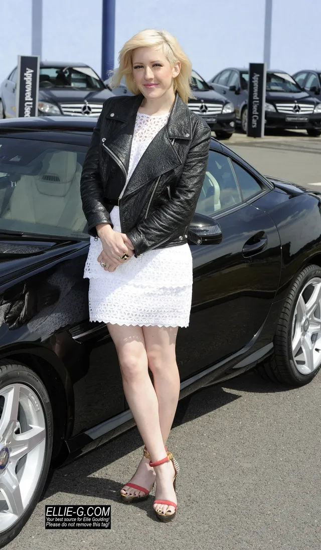Ellie Goulding posing beside a car in a white dress and pink heels.