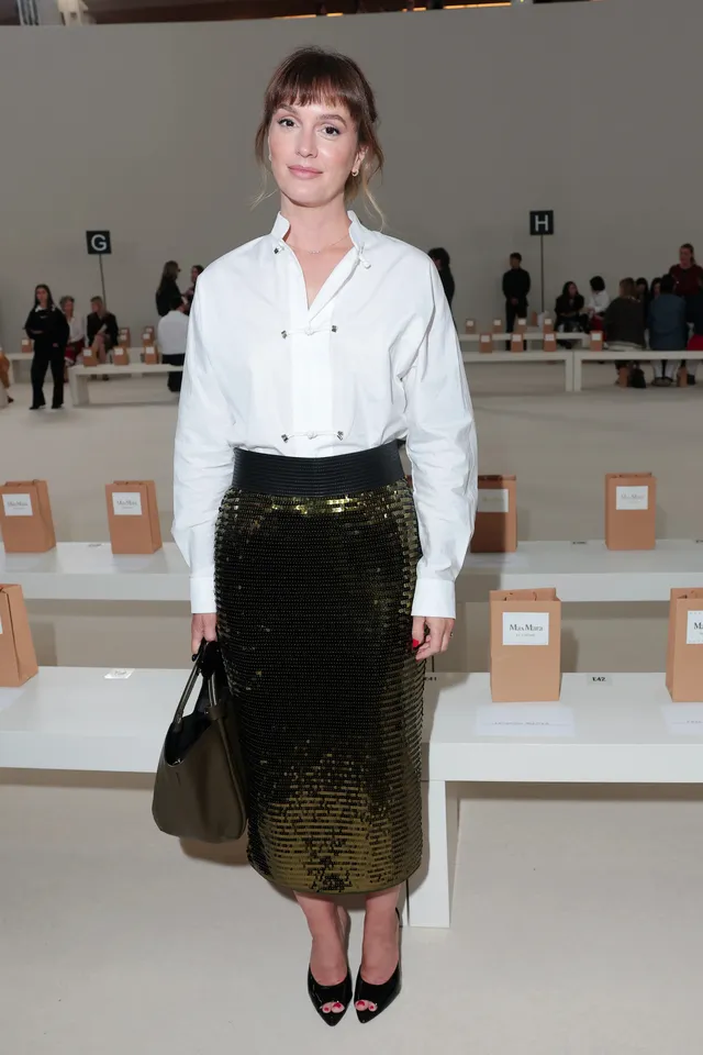 Leighton Meester posing at a fashion show in a white blouse and dark skirt.