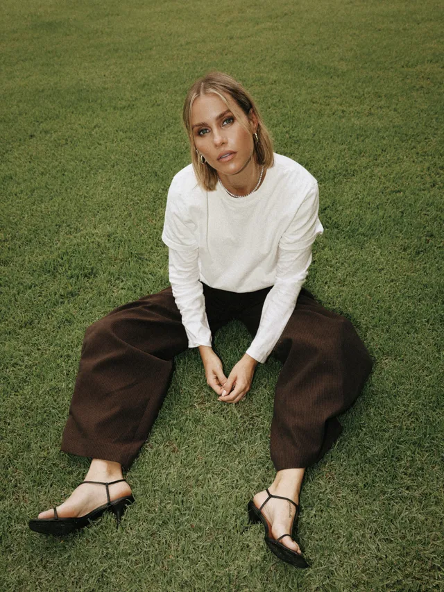 Claire Holt sitting on grass in a white top and brown trousers.