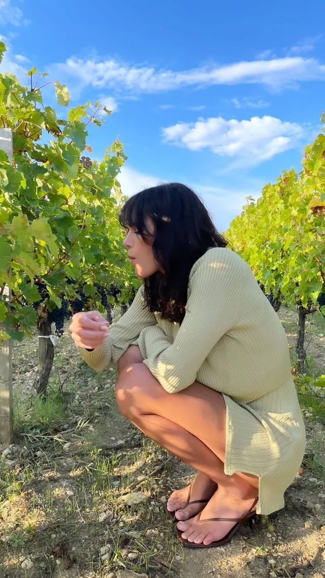 Bruna Marquezine crouching outdoors in a pale outfit and dark flip-flops.