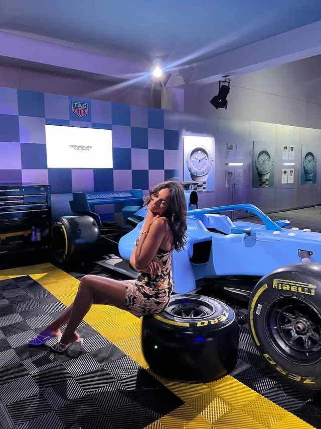 Greta Onieogou sitting beside a racing car display in a patterned dress and heels.