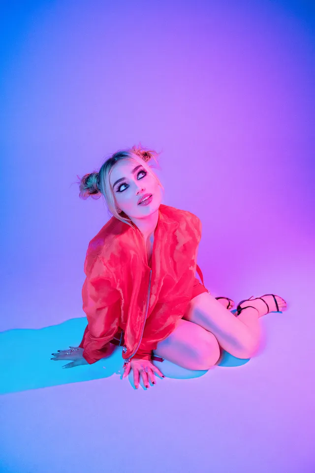 Meg Donnelly crouching in a red outfit and black heels for a colorful studio portrait.