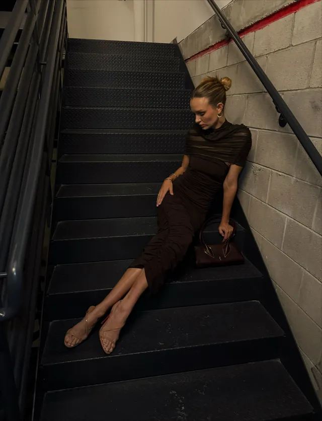 Josephine Skriver sitting on a staircase in a fitted dark dress and pointed heels.