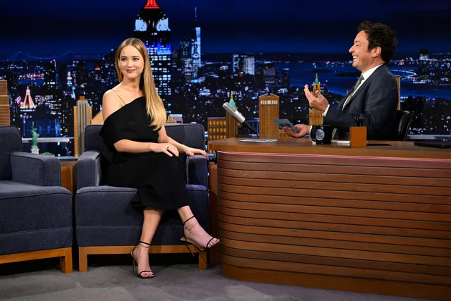 Jennifer Lawrence sitting during a television interview in a black dress and heels.